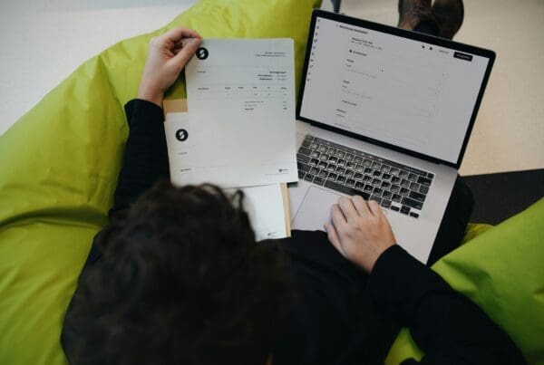 Personne assise sur un pouf vert consultant une facture sur papier et un formulaire sur un ordinateur portable