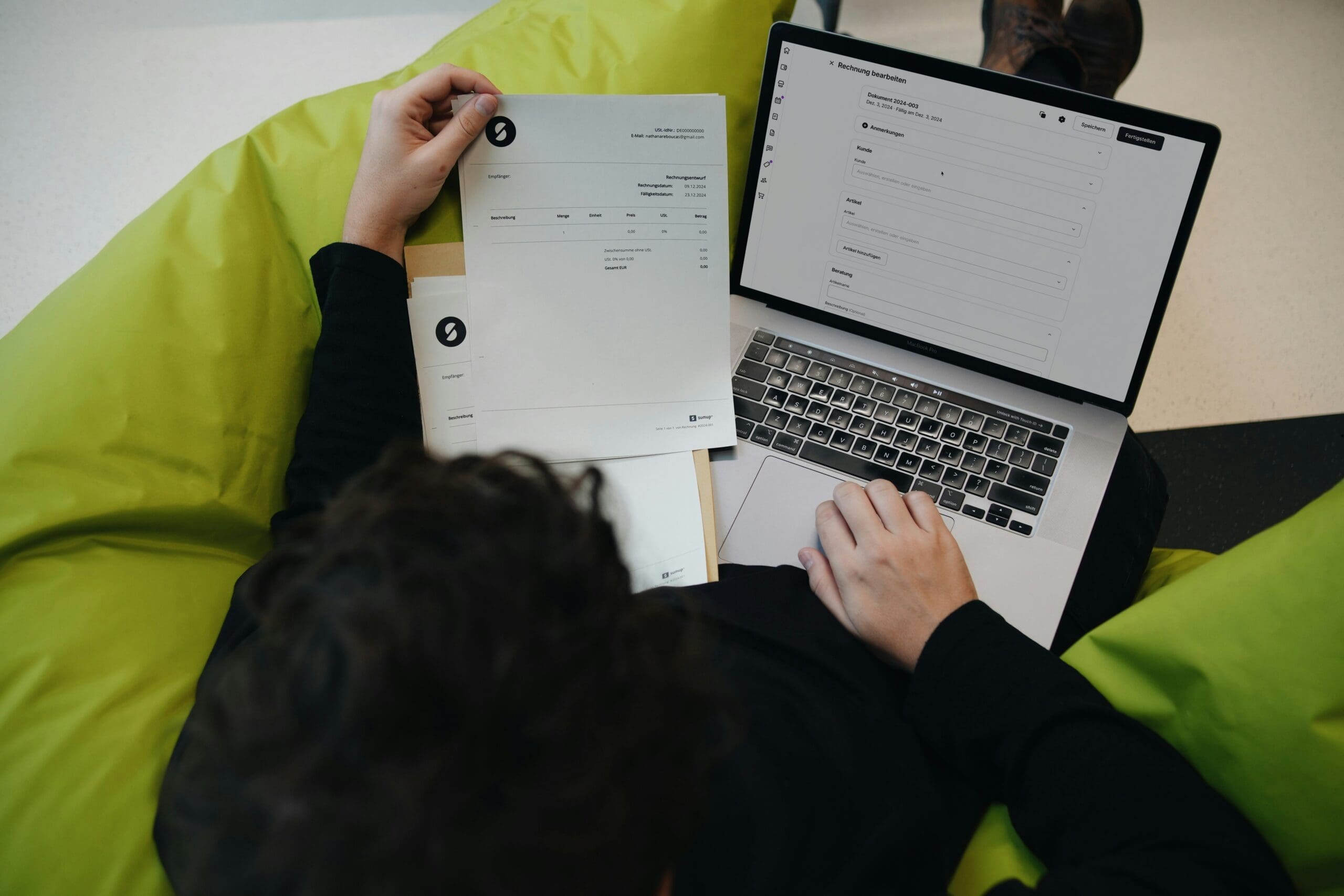 Personne assise sur un pouf vert consultant une facture sur papier et un formulaire sur un ordinateur portable